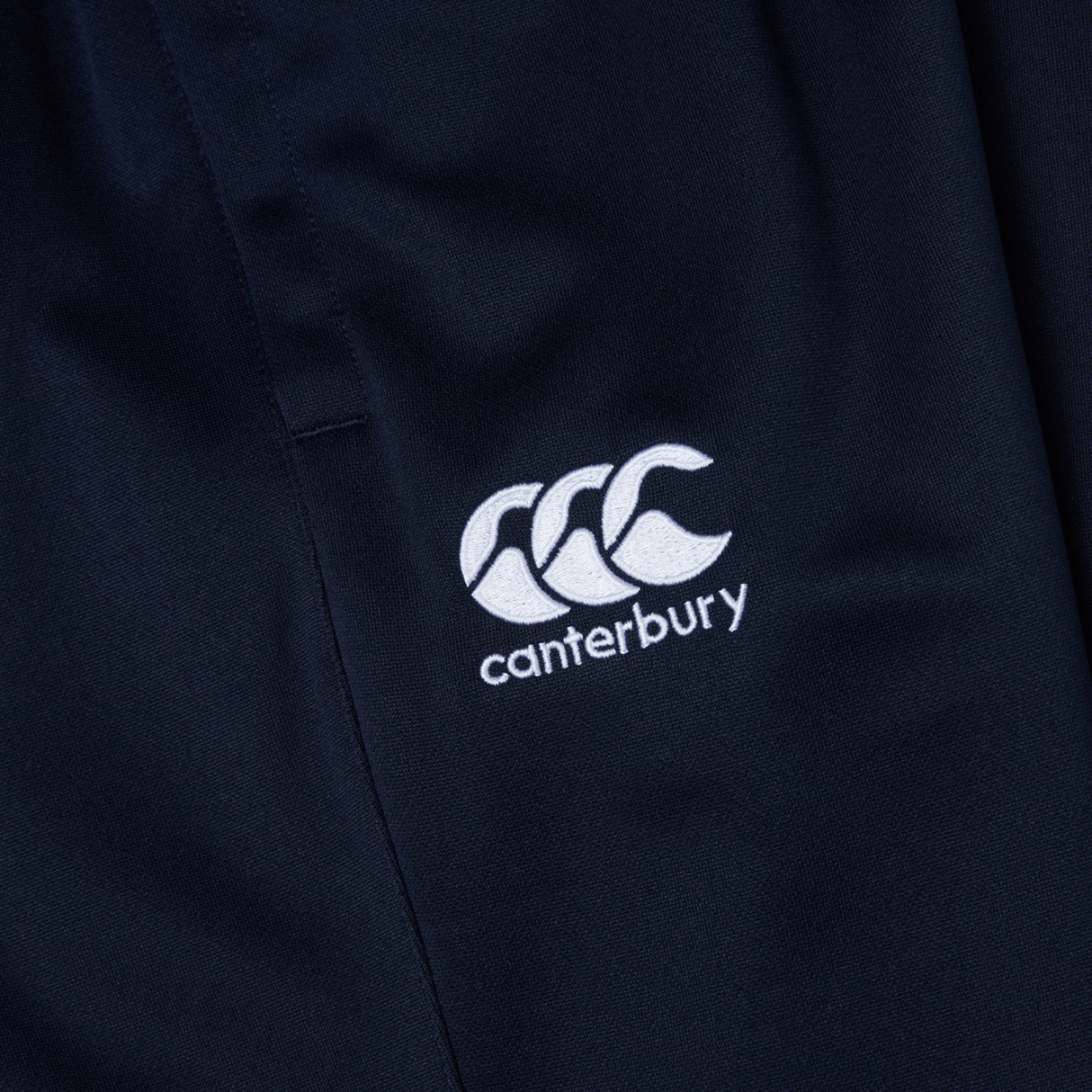 Close-up of navy blue fabric with a white embroidered logo of three overlapping figures and the text "Canterbury"—perfect for Canterbury Stretch Tapered Pants that are quick-dry and ideal for your workout.