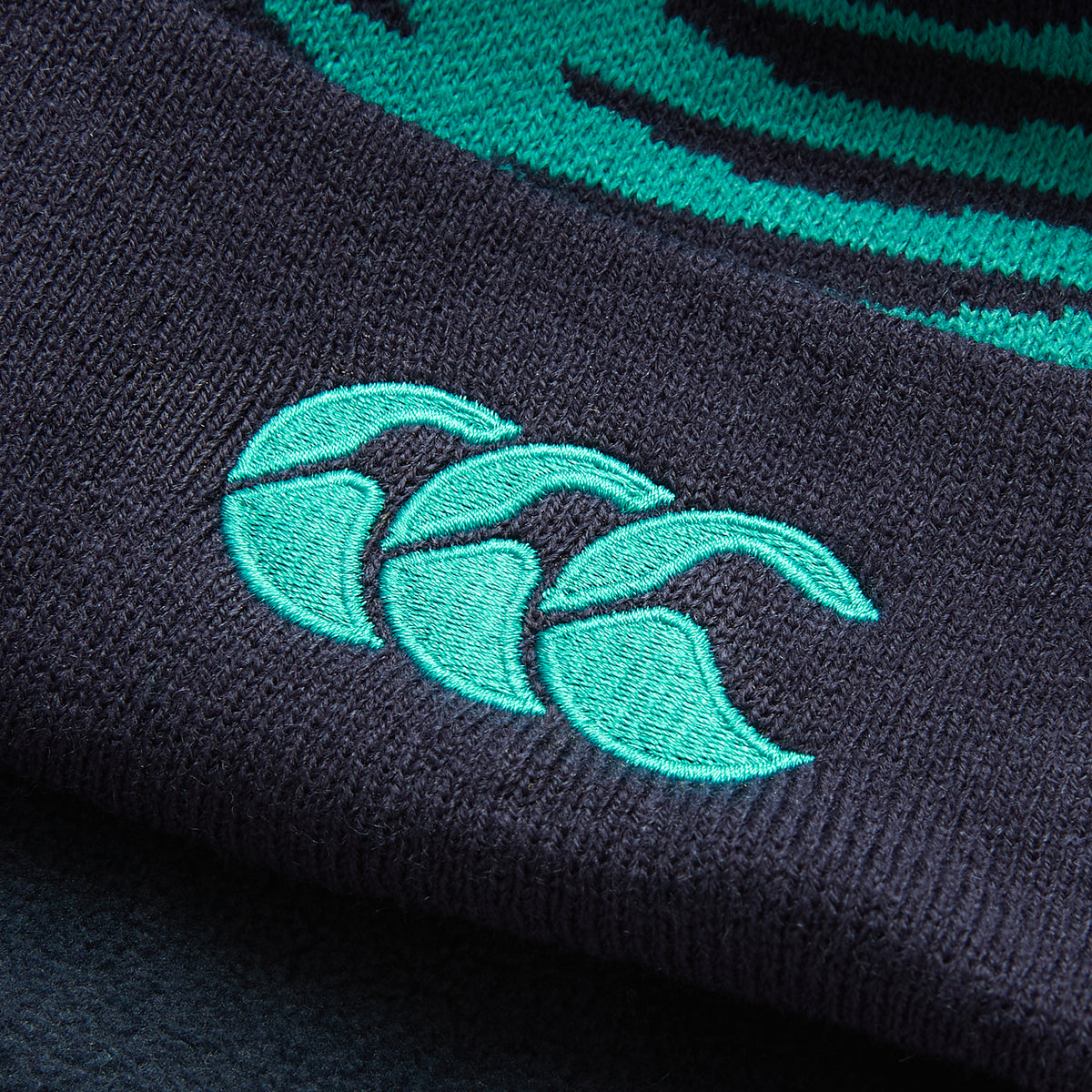 Close-up of a dark fabric featuring three teal overlapping crescent shapes embroidered in a stylized logo, reminiscent of the detailing on the Ireland 26 Bobble Hat by Canterbury.