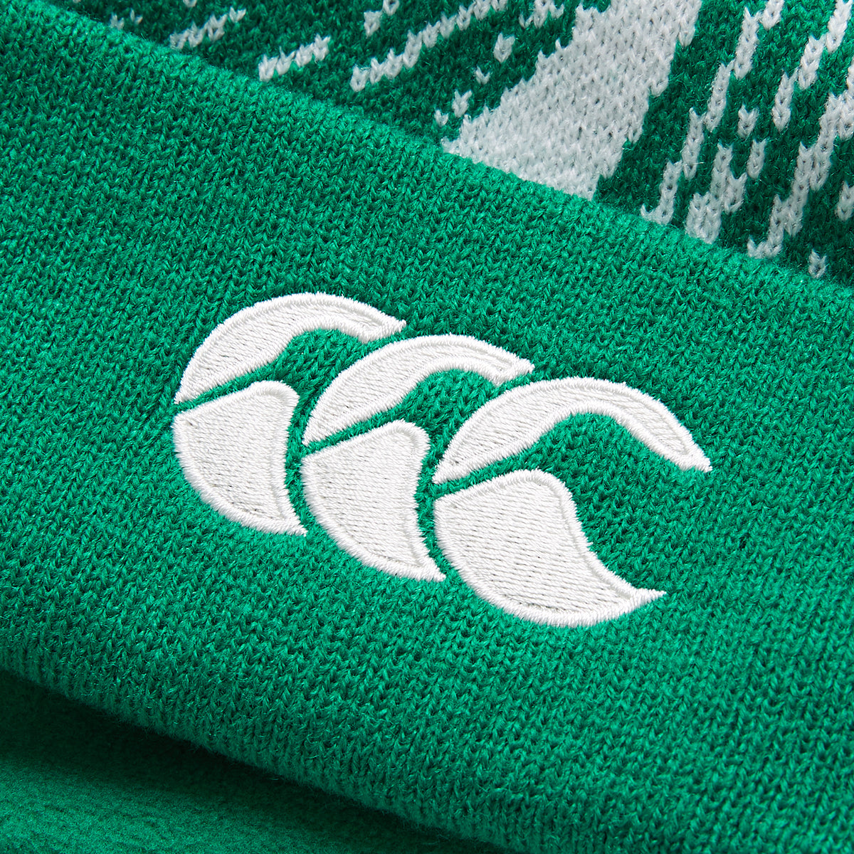 Close-up of a green knit fabric featuring an embroidered white logo with three stylized overlapping shapes, as seen on the Ireland 26 Bobble Hat by Canterbury.