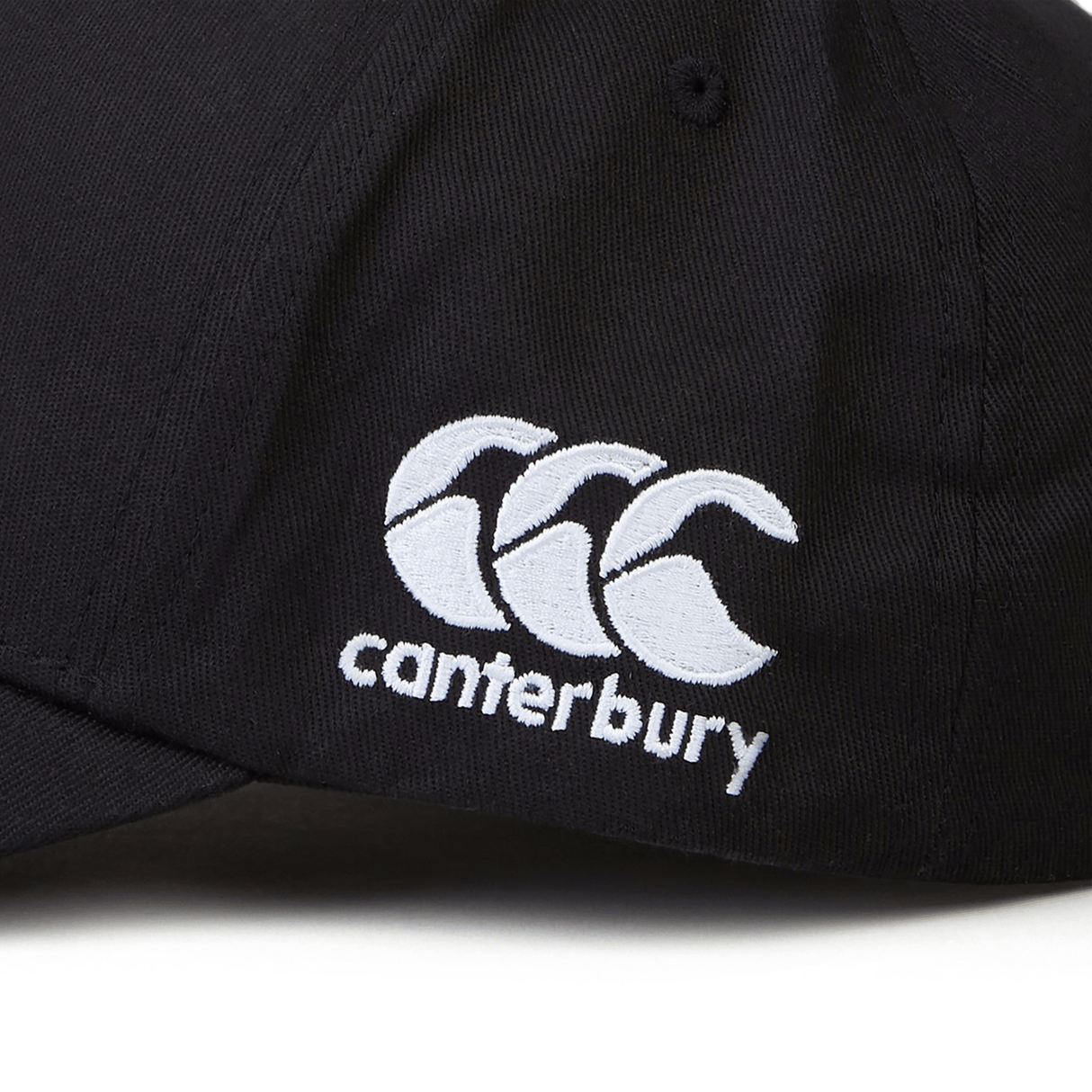 The Kenya Shield Flex Drill Cap by EMB Canterbury is a sleek black cap with an intricately embroidered white "canterbury" logo and three stylized "C" shapes on the front, combining elegance with adventure spirit.