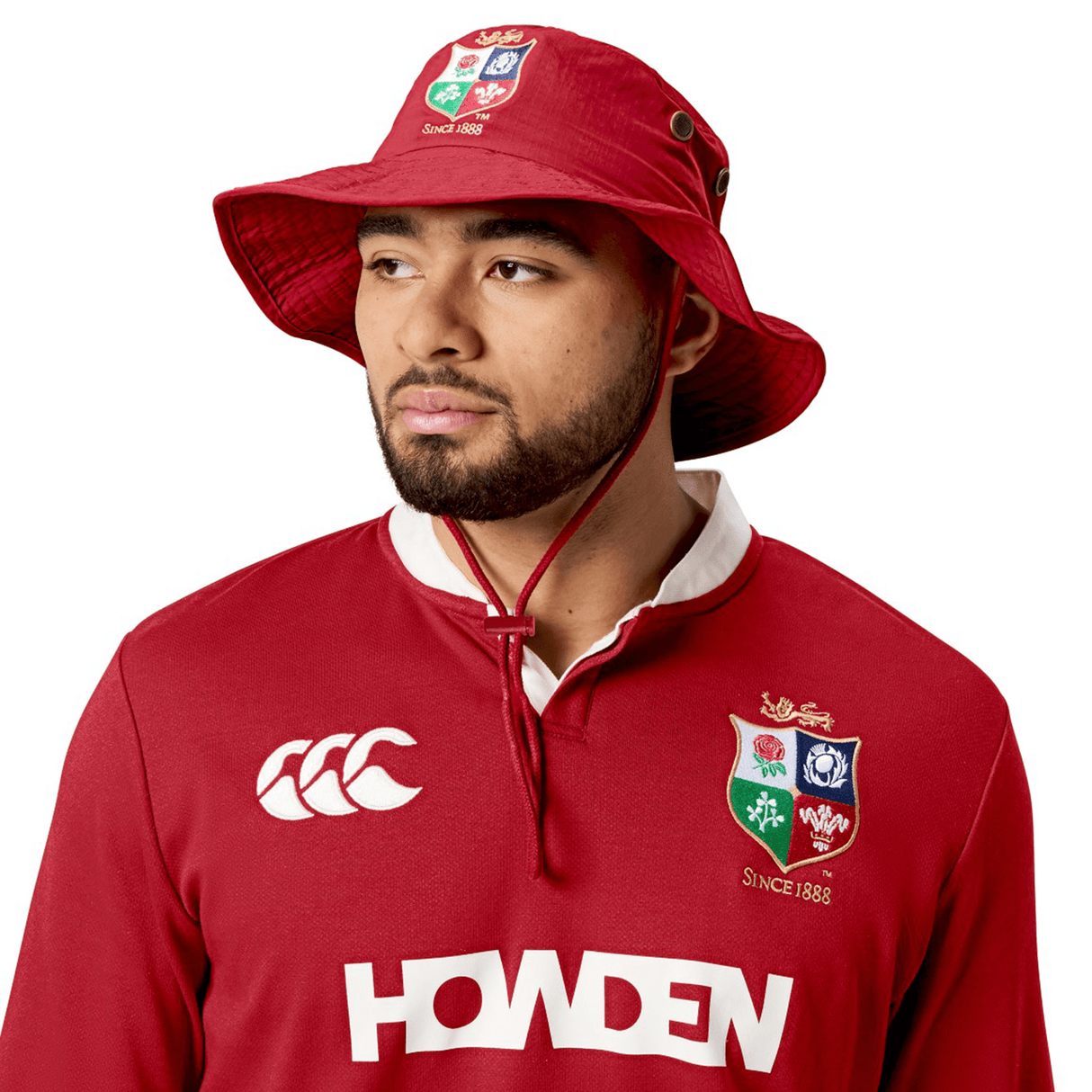 A person wearing a red rugby jersey adorned with sponsor logos pairs it with the British & Irish Lions 2025 Wide Brim Bucket Hat by Canterbury, crafted from quick-dry ripstop fabric, and looks to the side.