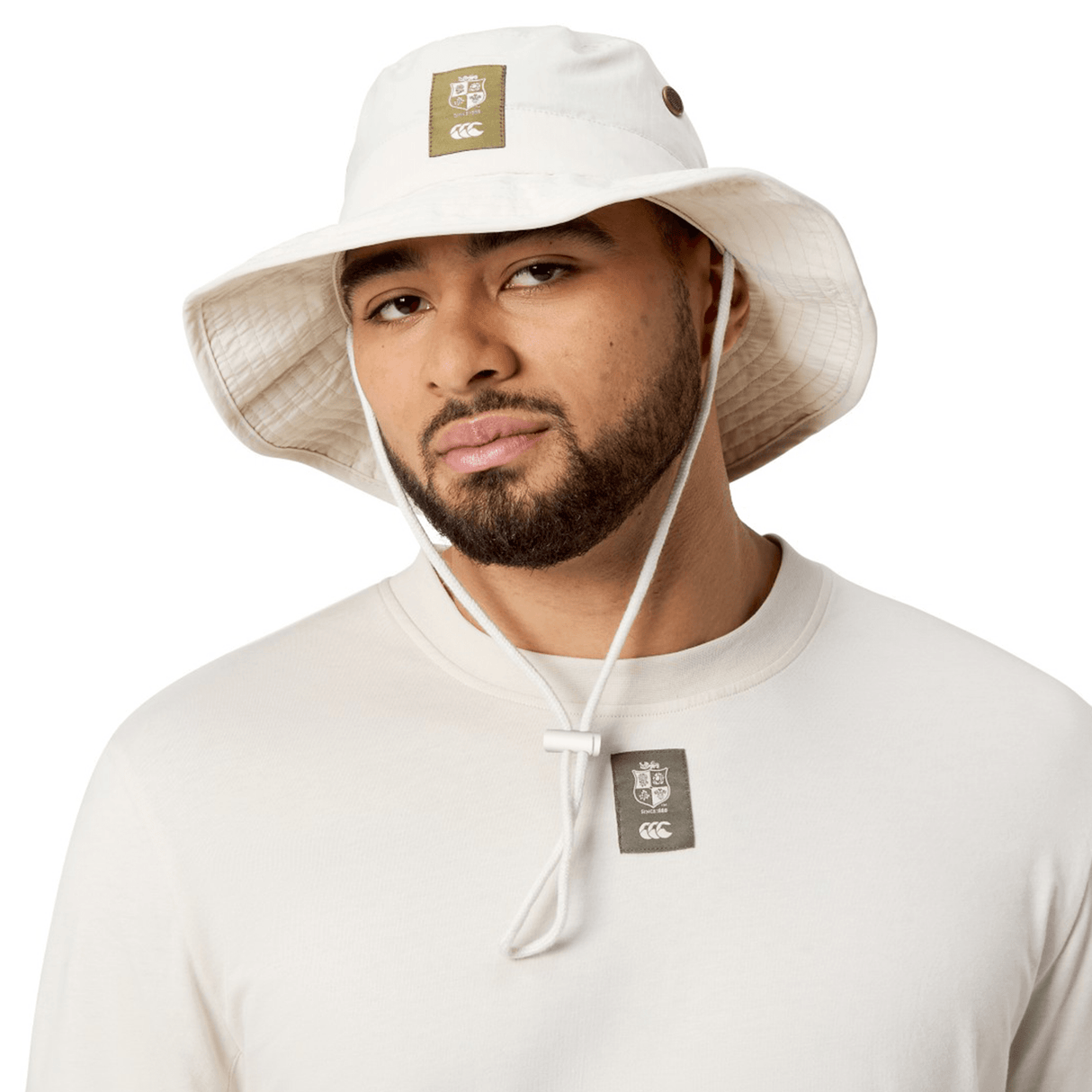A person wearing the British & Irish Lions 2025 Outback Wide Brim Bucket Hat by Canterbury, made of white quick-dry ripstop fabric, and a long-sleeve shirt with a logo tag, looking at the camera.