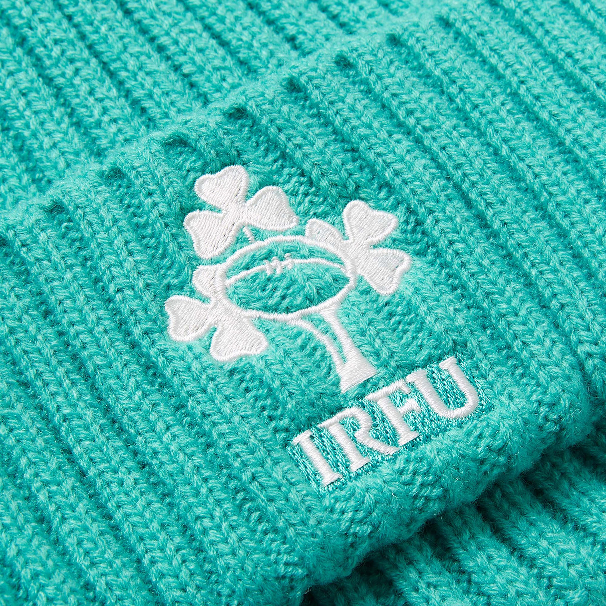 Close-up of a turquoise knit fabric with a white IRFU logo featuring three shamrocks and a rugby ball, as seen on the Ireland 26 Beanie by Canterbury.