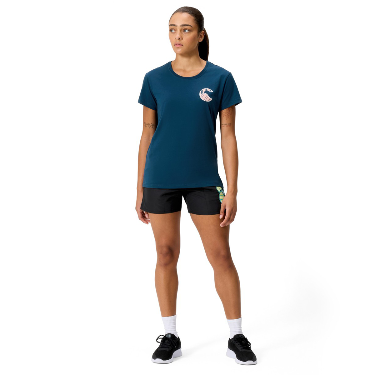 A person stands facing forward in a Canterbury Women's Uglies Logo T-Shirt, paired with black shorts, white socks, and black sneakers against a plain white background.