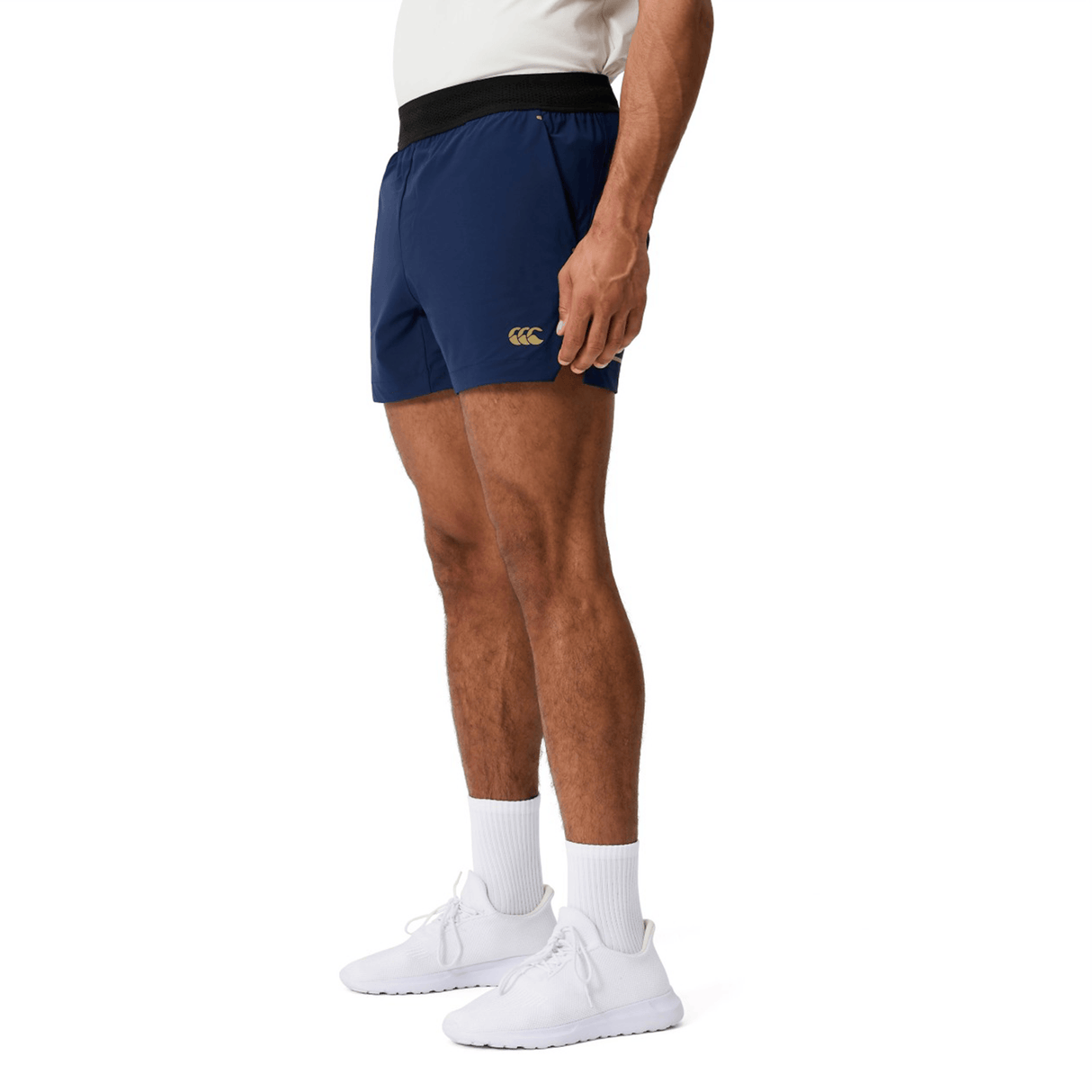 Man in a white shirt, Canterbury Everest Woven Short 5" in navy with logo, white calf socks, and white sneakers stands against a plain background. These Canterbury shorts offer breathable wicking fabric for all-day comfort.