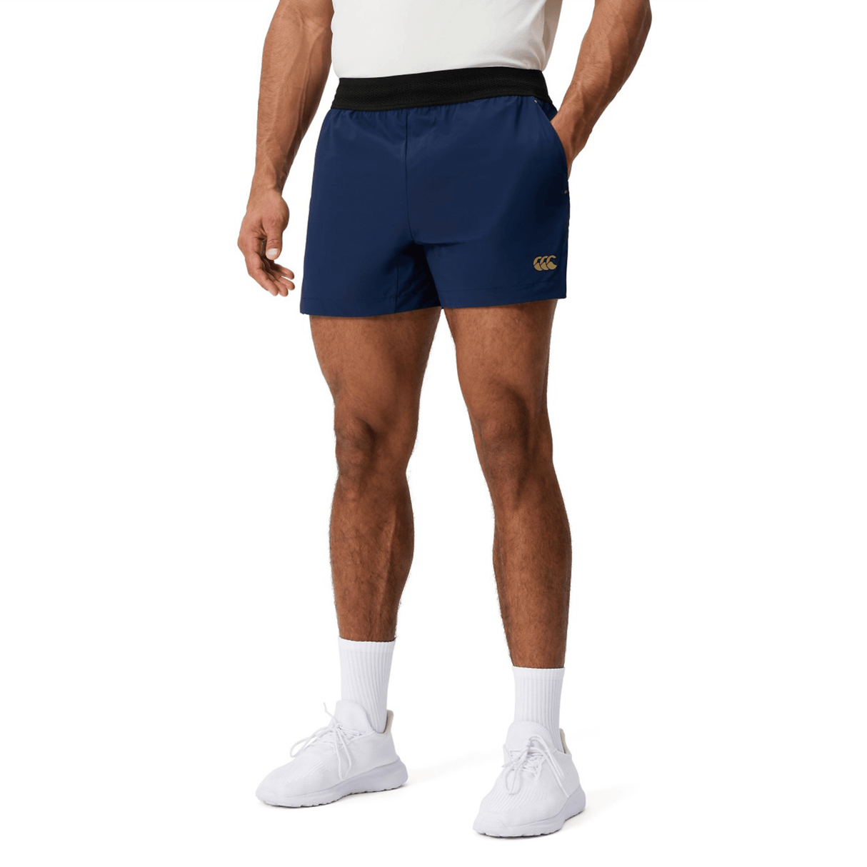 A person wearing a white shirt, Canterbury Everest Woven Short 5" gym shorts by Canterbury, white socks, and white sneakers stands against a plain background. The navy blue shorts are made of breathable wicking fabric.