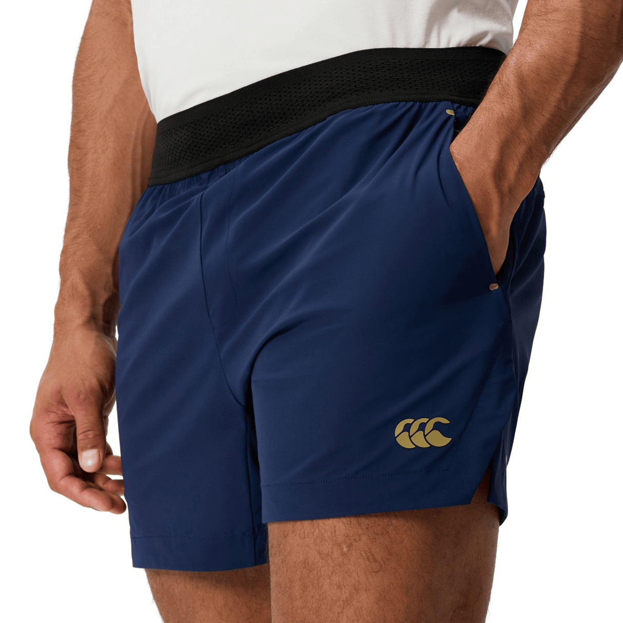 A person wears a white shirt and Canterbury Everest Woven Short 5", navy blue with a black waistband, gold Canterbury logo on the left leg, and made from breathable wicking fabric for comfort.