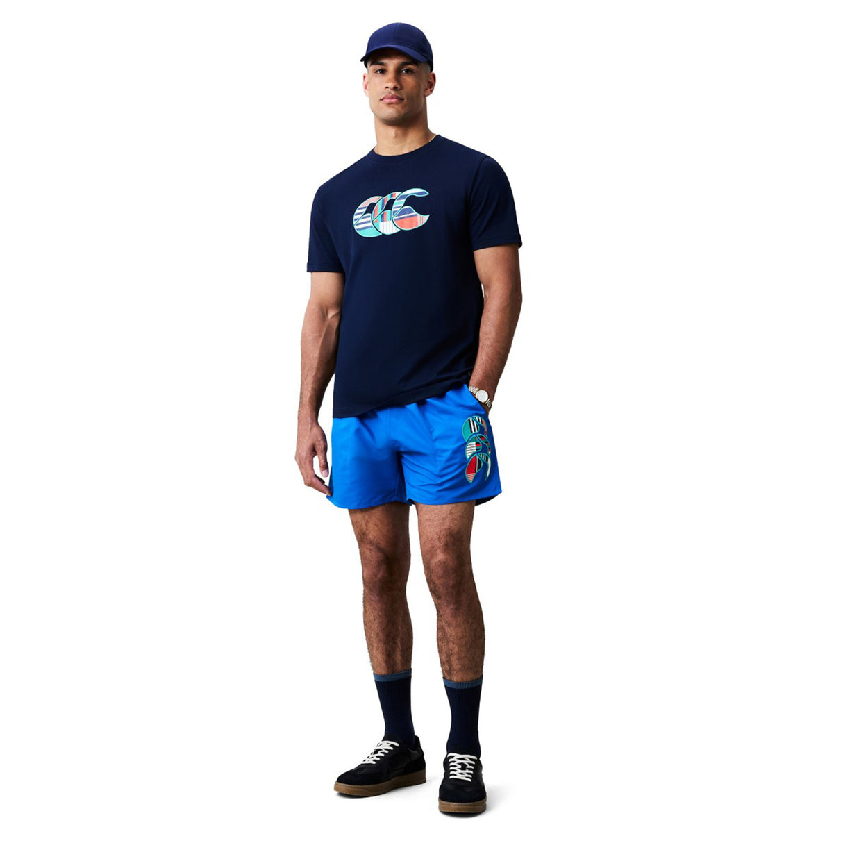 A man stands against a plain white background wearing a navy cap, navy graphic t-shirt, Canterbury Uglies Tactic Short by Canterbury with logo, navy socks, and black sneakers.