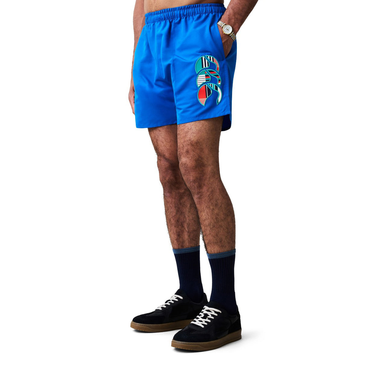 Man wearing Canterbury Uglies Tactic Short by Canterbury, which features a bright blue design with a colorful lobster graphic, paired with dark socks, black sneakers, and a watch, standing against a white background.