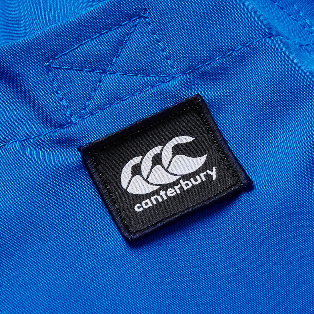 A close-up of blue fabric with a black and white Canterbury logo patch stitched on, highlighting the iconic detail of Canterbury Uglies Tactic Short—rugby training shorts from Canterbury.