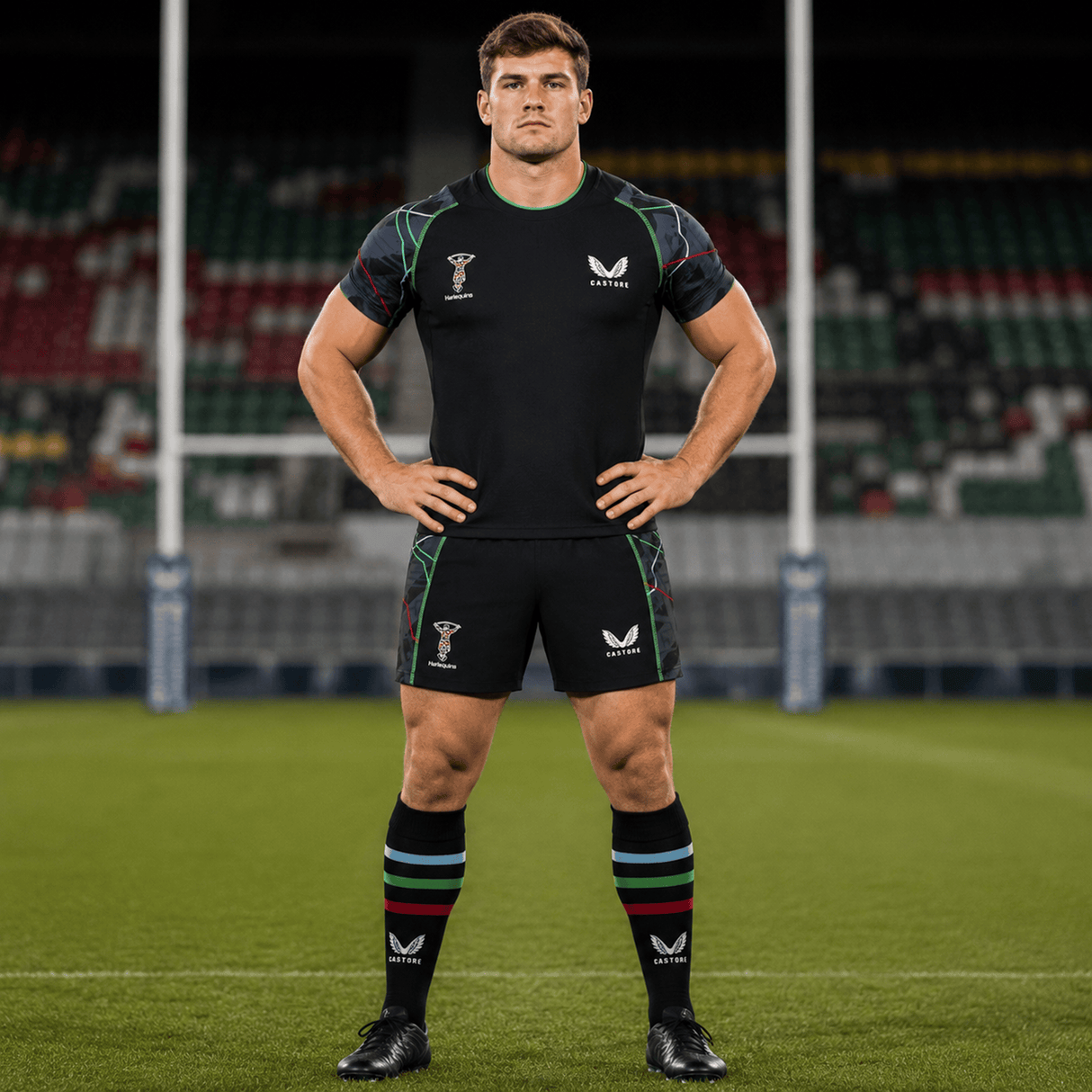 A male rugby player in a black Castore kit stands hands on hips on a rugby field, wearing Harlequins 22/23 3rd Pro Short by Castore, with goalposts and empty stadium seats behind him.