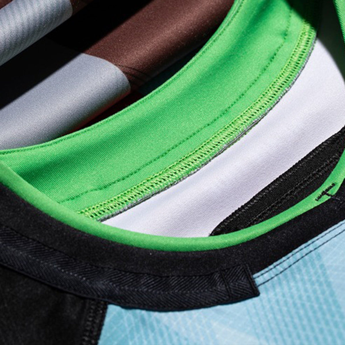 Close-up of overlapping rugby jerseys, highlighting the collars and stitching of the Castore Harlequins 23/24 Pro Home Jersey in green, black, white, and light blue fabric.