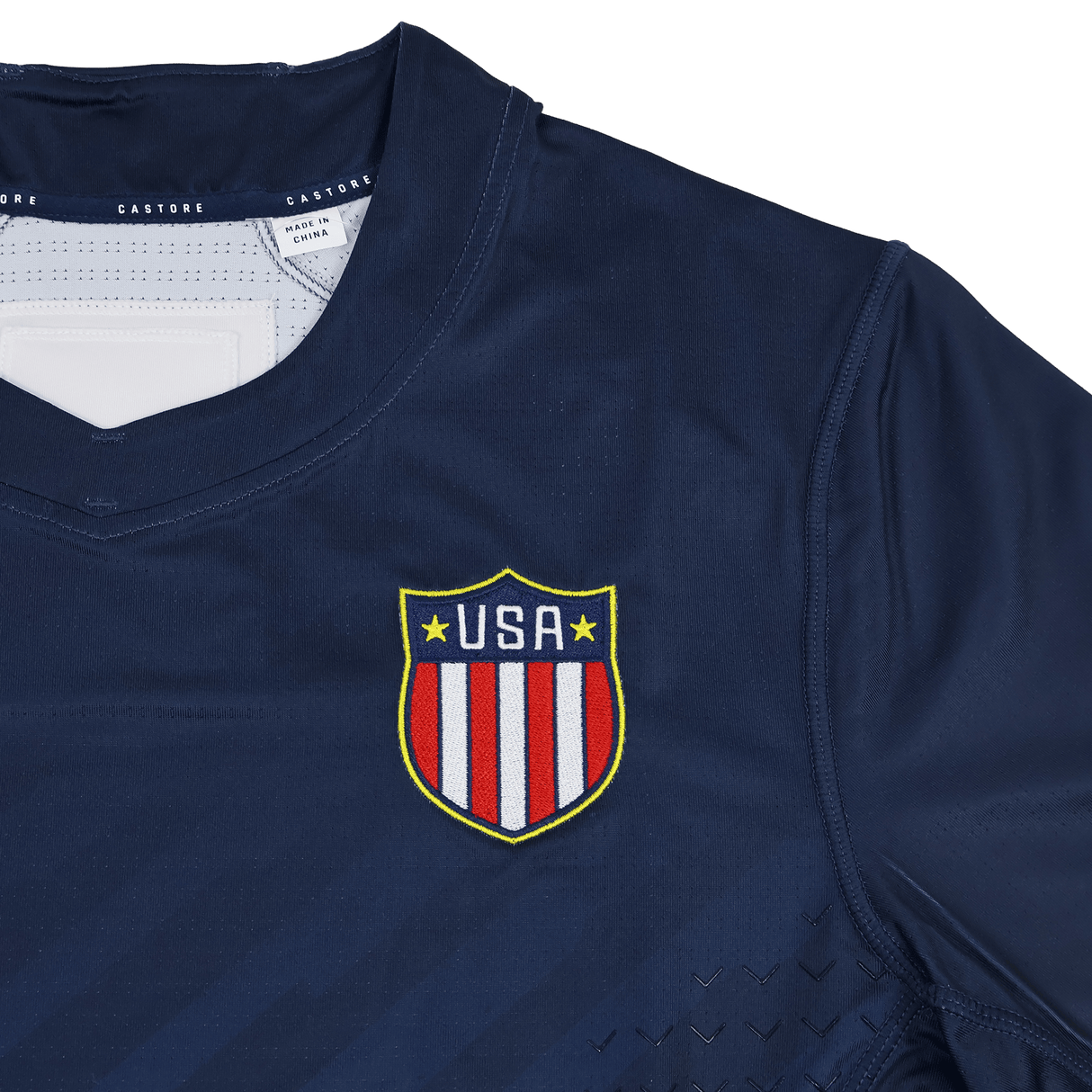 The USA Rugby 24/25 7s Olympic Pro Home Jersey by Castore features a navy blue design with a shield emblem of red and white stripes and yellow stars on the chest, inspired by Olympic style.