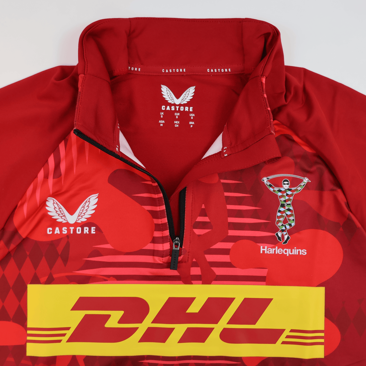 The Harlequins 24/25 Matchday 1/4 Zip Midlayer by Castore features a DHL logo, partial zipper, and a multicolor Harlequin figure on the right chest.