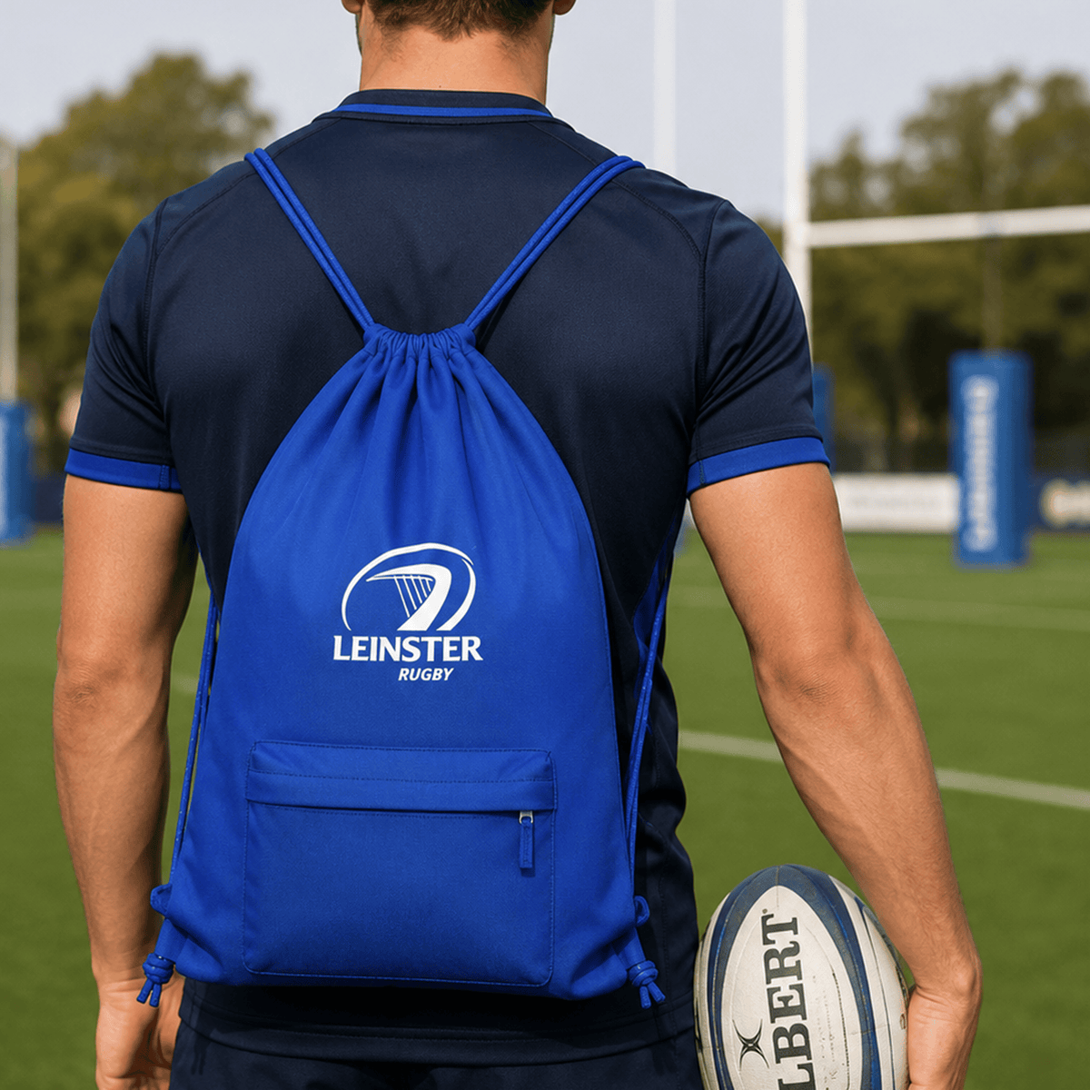 A person in navy sportswear stands on a rugby field holding a rugby ball and wearing the Castore Leinster 24/25 Drawstring Bag, featuring the Leinster Rugby logo.