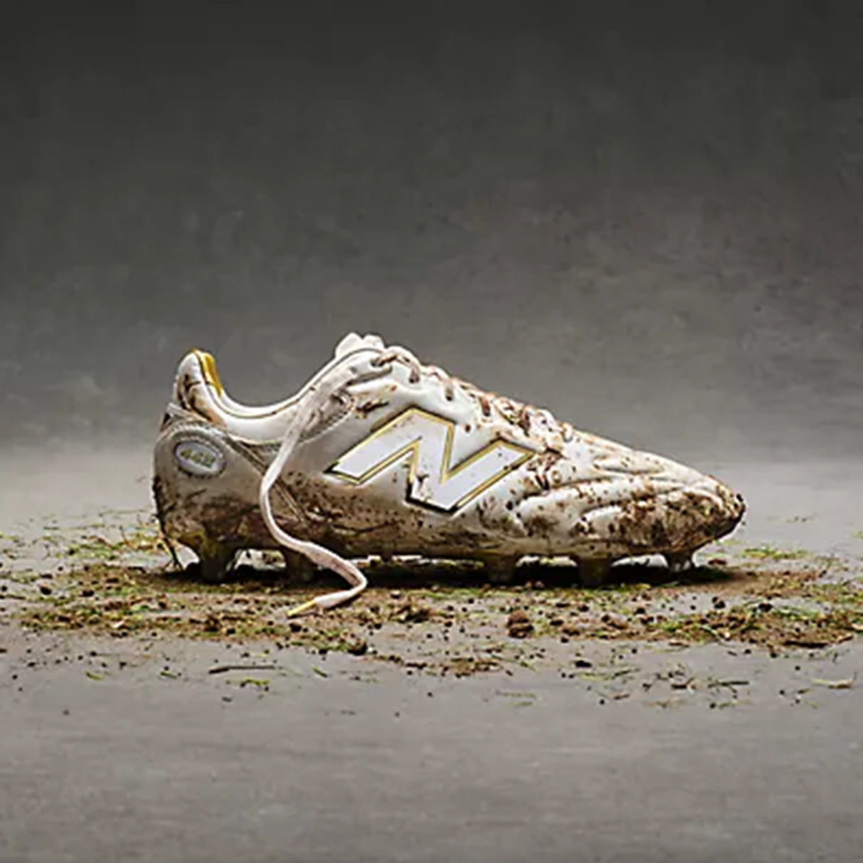 A single New Balance 442 Elite FG v3 cleat in Sea Salt/Gold Metallic is coated in mud and grass, resting on a gray surface with scattered dirt and debris.