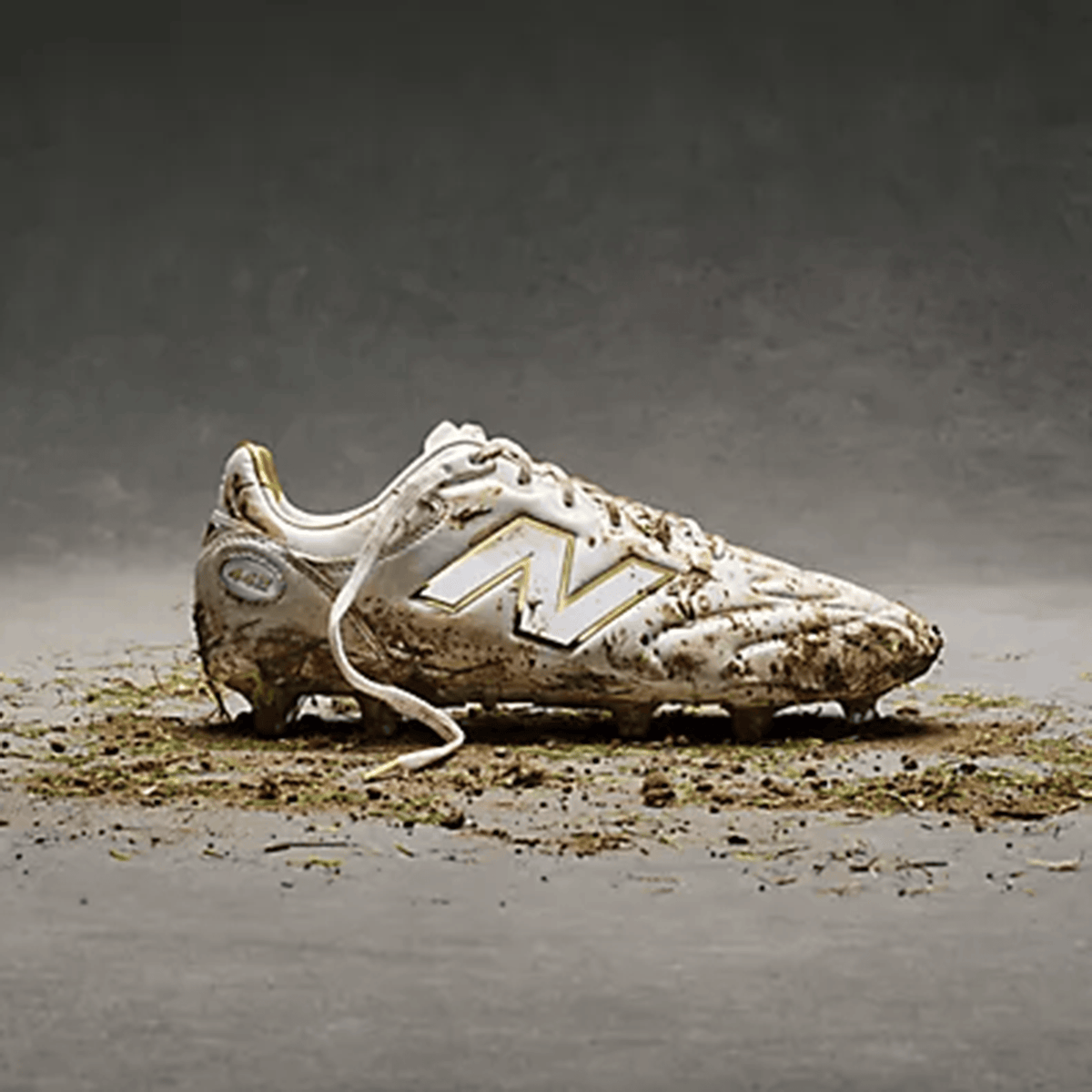 A single New Balance 442 Elite FG v3 cleat in Sea Salt/Gold Metallic is coated in mud and grass, resting on a gray surface with scattered dirt and debris.