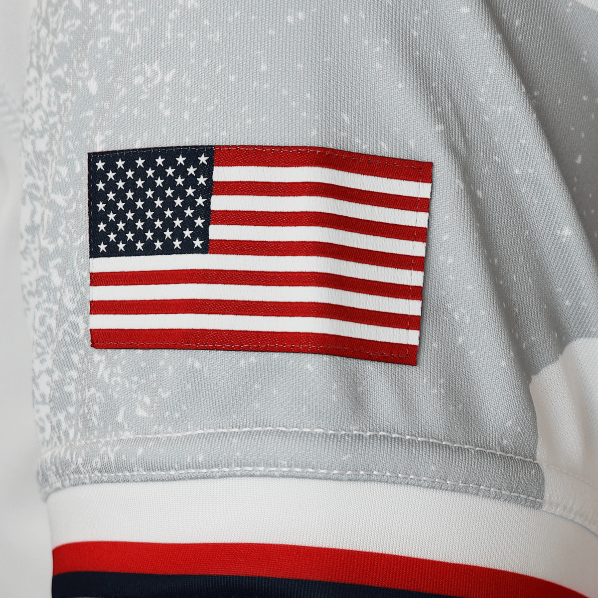 A close-up of a gray fabric sleeve with an embroidered United States flag patch, part of the Women's USA Rugby WRWC25 Replica Away Jersey by Macron.