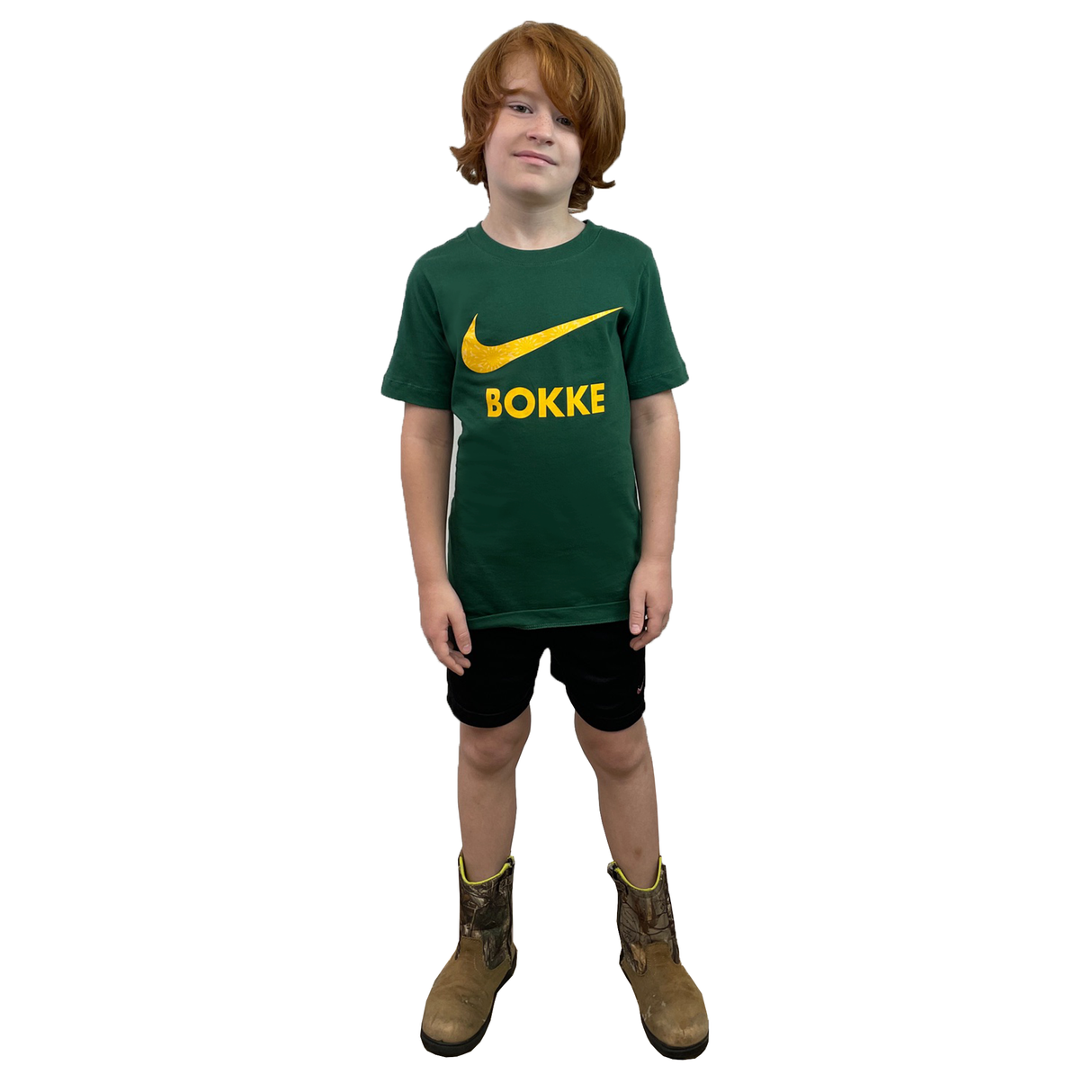 A young boy with red hair stands facing forward on a white background, wearing the Nike Youth Springboks 25 Bokke T-Shirt, paired with black shorts and brown work boots.