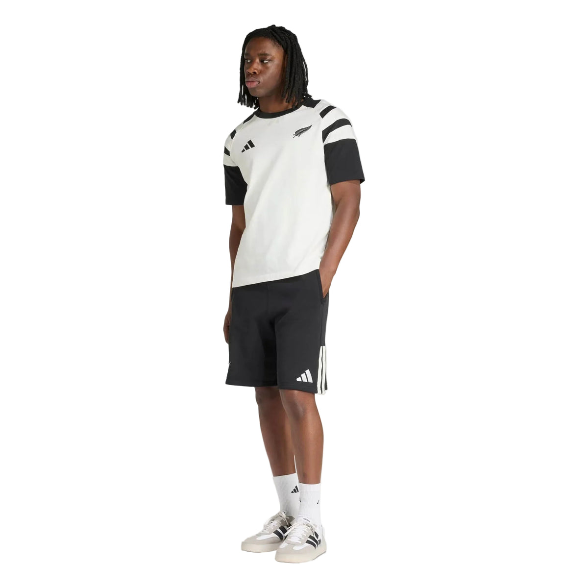 Person wearing a white and black T-shirt, adidas All Blacks 26/27 Leisurewear Shorts in black with white stripes, white socks, and white sneakers, standing against a plain white background.