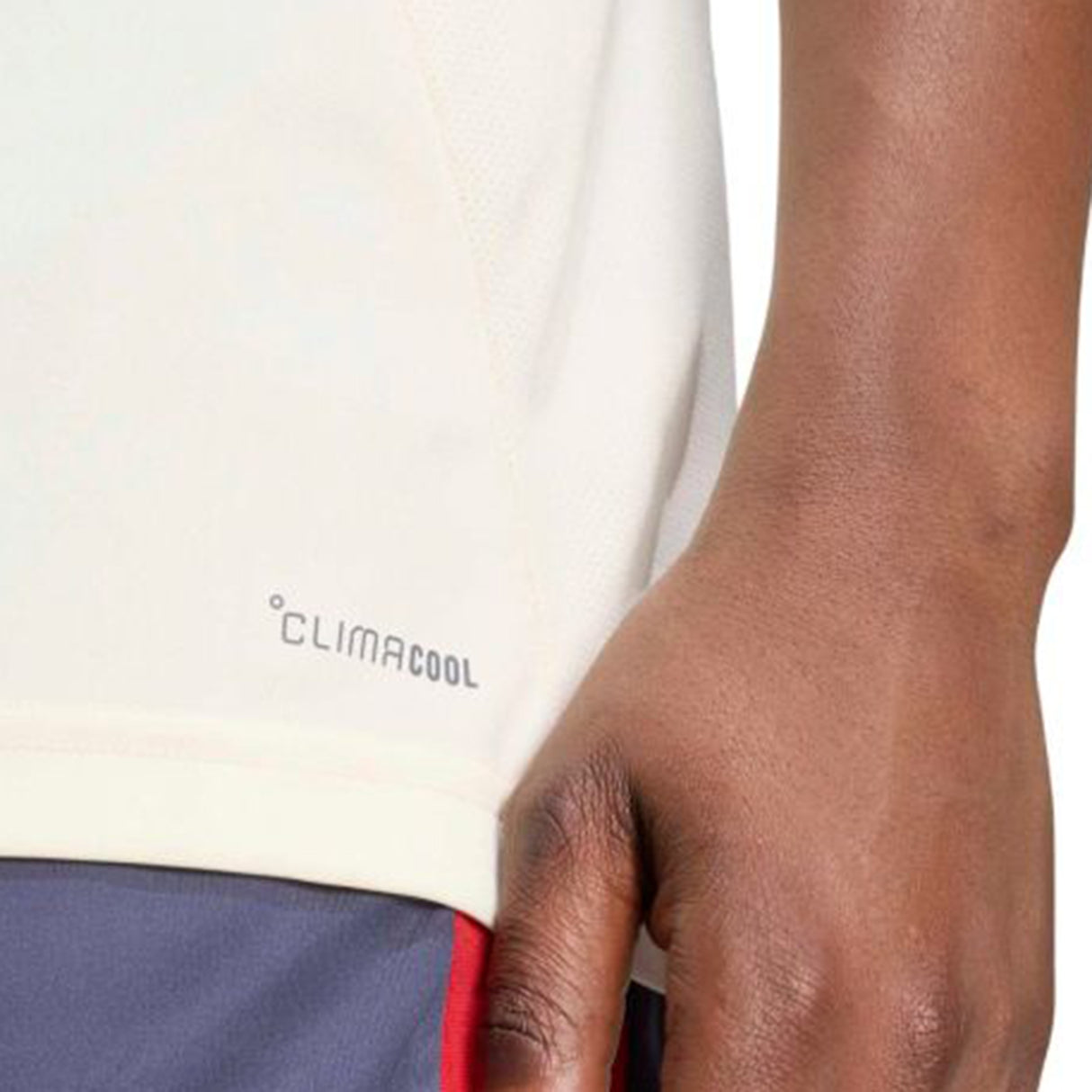 Close-up of a person’s hand resting by their side, revealing part of a white France FFR 26/27 Performance T-Shirt by adidas with “CLIMACOOL” labeling, paired with blue shorts featuring a red stripe—evoking the look of the French national rugby team.