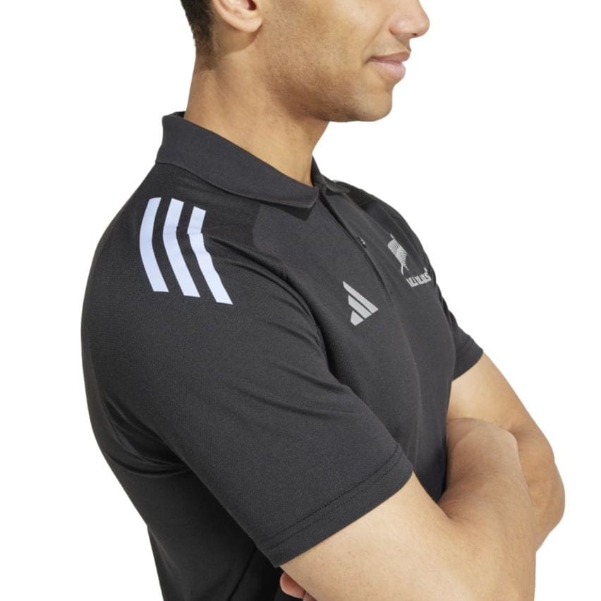 Dressed in an adidas All Blacks 24/25 Performance Polo, a person stands with arms crossed and facing sideways.