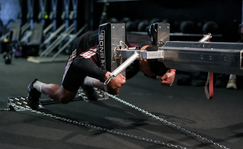 A person in a black and white athletic outfit performs a rowing motion with a chain-pulled ergometer in a dark gym setting.