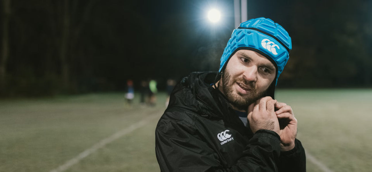 A man in a black sports jacket adjusts his ear while wearing a blue padded rugby helmet on a lit outdoor field at night.
