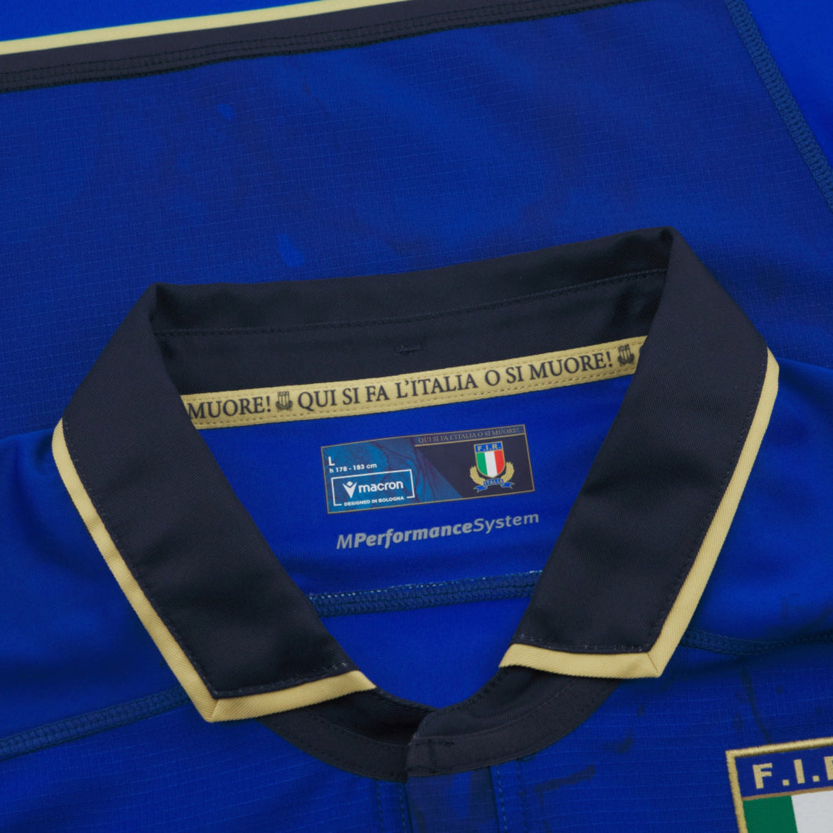 Close-up of the Italy 25/26 Authentic Home Jersey by Macron shows the blue collar with gold trim, Italian flag, Macron logo, and "Italy rugby jersey" text printed inside the neckline.