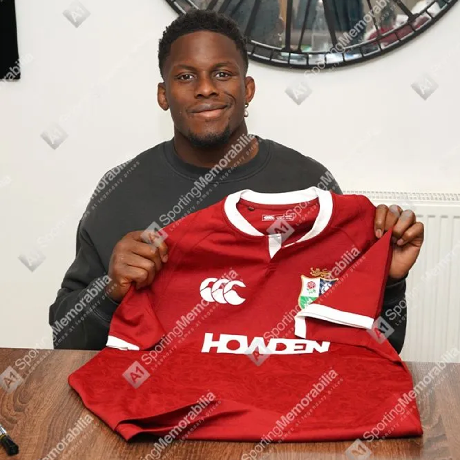 A man sits indoors holding a Maro Itoje Front Signed British & Irish Lions 2025 Shirt by Icons—a red rugby jersey with white trim—displayed as prized memorabilia in front of a radiator and circular mirror.