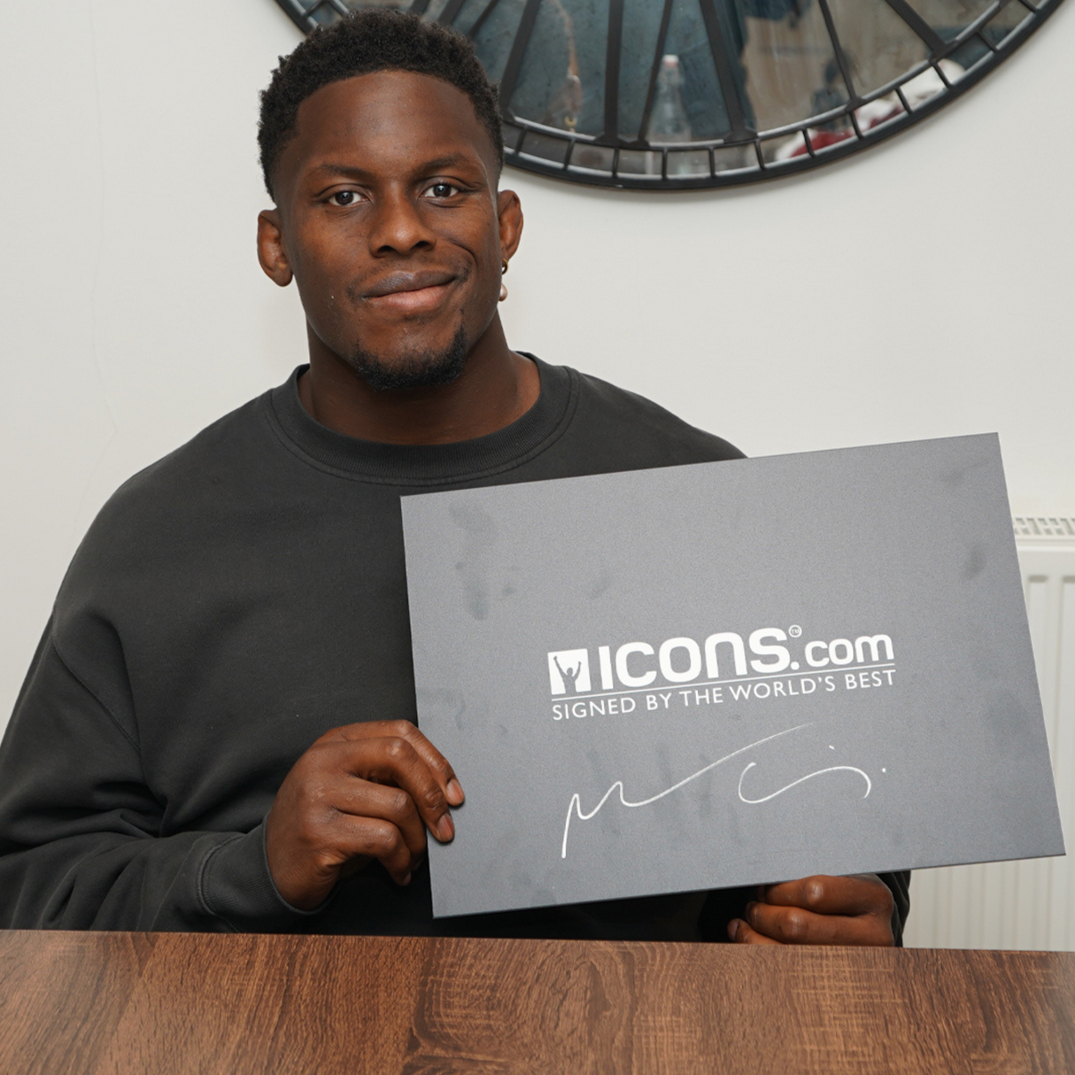 A man in a dark sweatshirt sits at a table holding an "ICONS.com SIGNED BY THE WORLD'S BEST" sign, next to an Icons Maro Itoje Signed British & Irish Lions Rugby Ball.