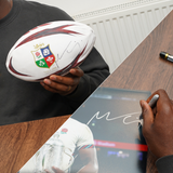 A person holds an Icons Maro Itoje Signed British & Irish Lions Rugby Ball and signs a photograph at a table with pens nearby, creating unique autographed rugby memorabilia.