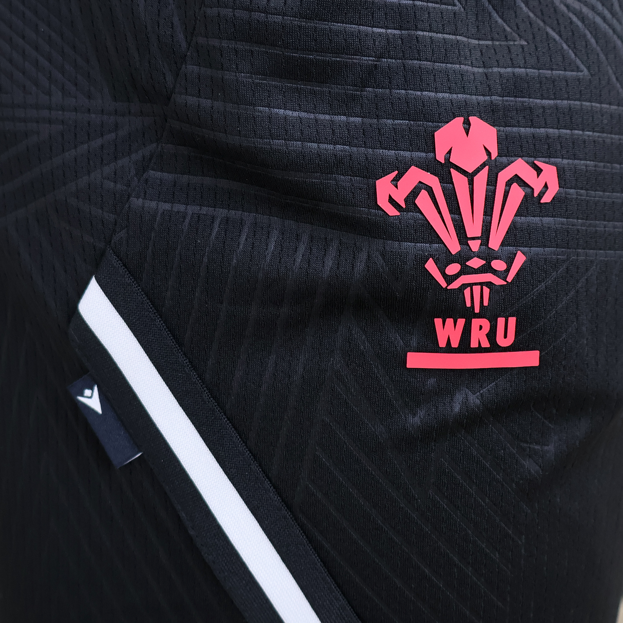 A close-up of the Macron Wales 25/26 Basketball Shorts, black with red and white stripes, showcasing the Welsh Rugby Union (WRU) logo for a modern athletic look.
