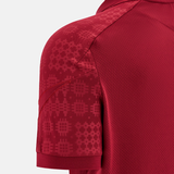 Close-up of the Wales 25/26 Player Travel Poly Polo by Macron, a red short-sleeve athletic shirt with a subtle geometric pattern on the sleeve and shoulder, inspired by player travel polos, shown on a plain background.