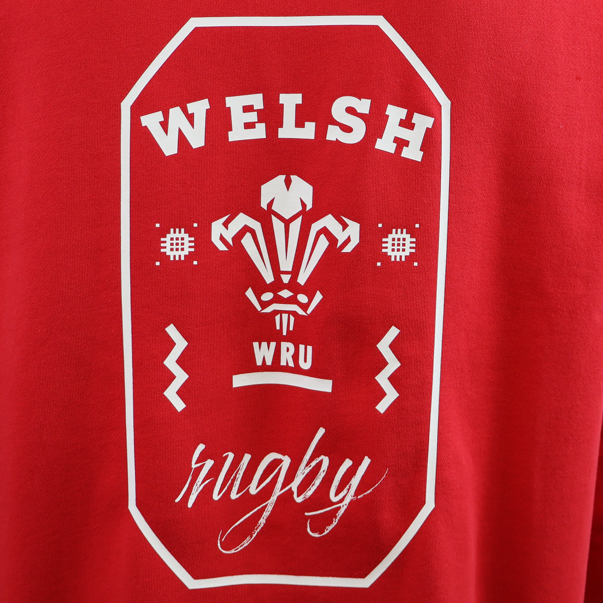 Red shirt with a white “Welsh rugby” print, WRU logo, and decorative elements—ideal for pairing with your Wales 25/26 Welsh Rugby Cotton Fan Hoodie by Macron.