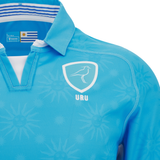 A close-up of the Uruguay URU 25/26 Replica Home Jersey by Macron shows light blue fabric, a bird emblem, "URU" text, sun patterns, and an Uruguayan flag tag inside the collar.
