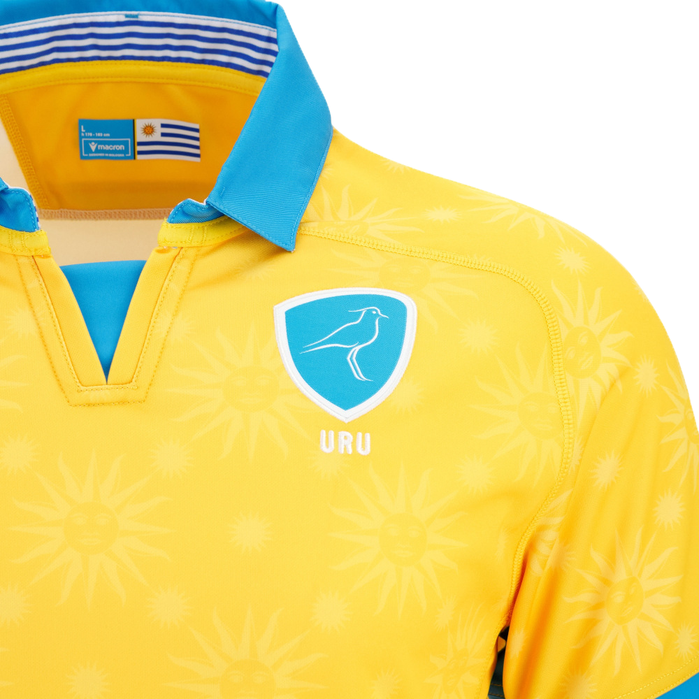 The Uruguay URU 25/26 Replica Away Jersey by Macron features a yellow design with blue collar, bird emblem, "URU" text, sun motifs, Uruguay flag tag inside the neck, and EcoFightex fabric. Official away jersey by Macron.