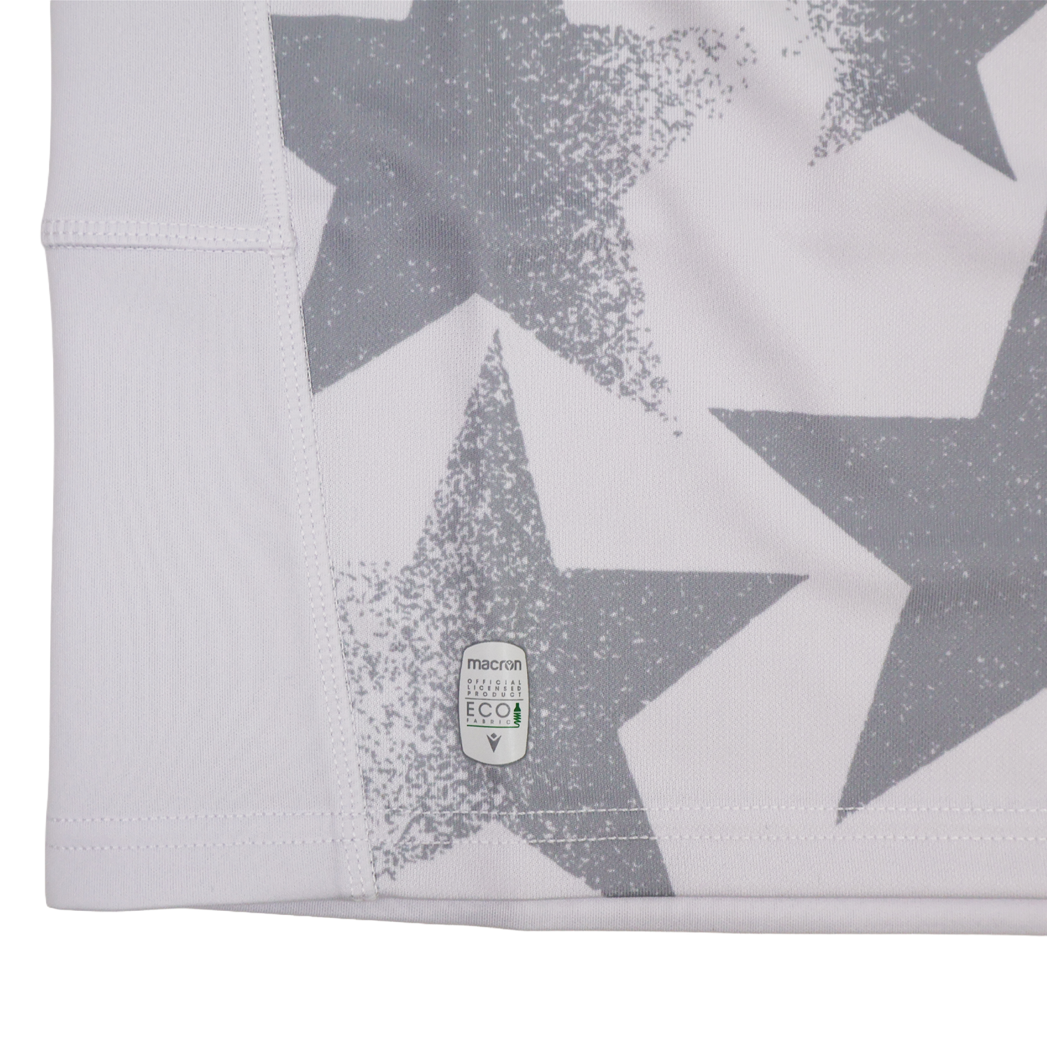 Close-up of a white fabric with large gray stars and a small "macron ECO" label near the hem, from the Youth USA Rugby WRWC 25 Home Jersey by Macron.