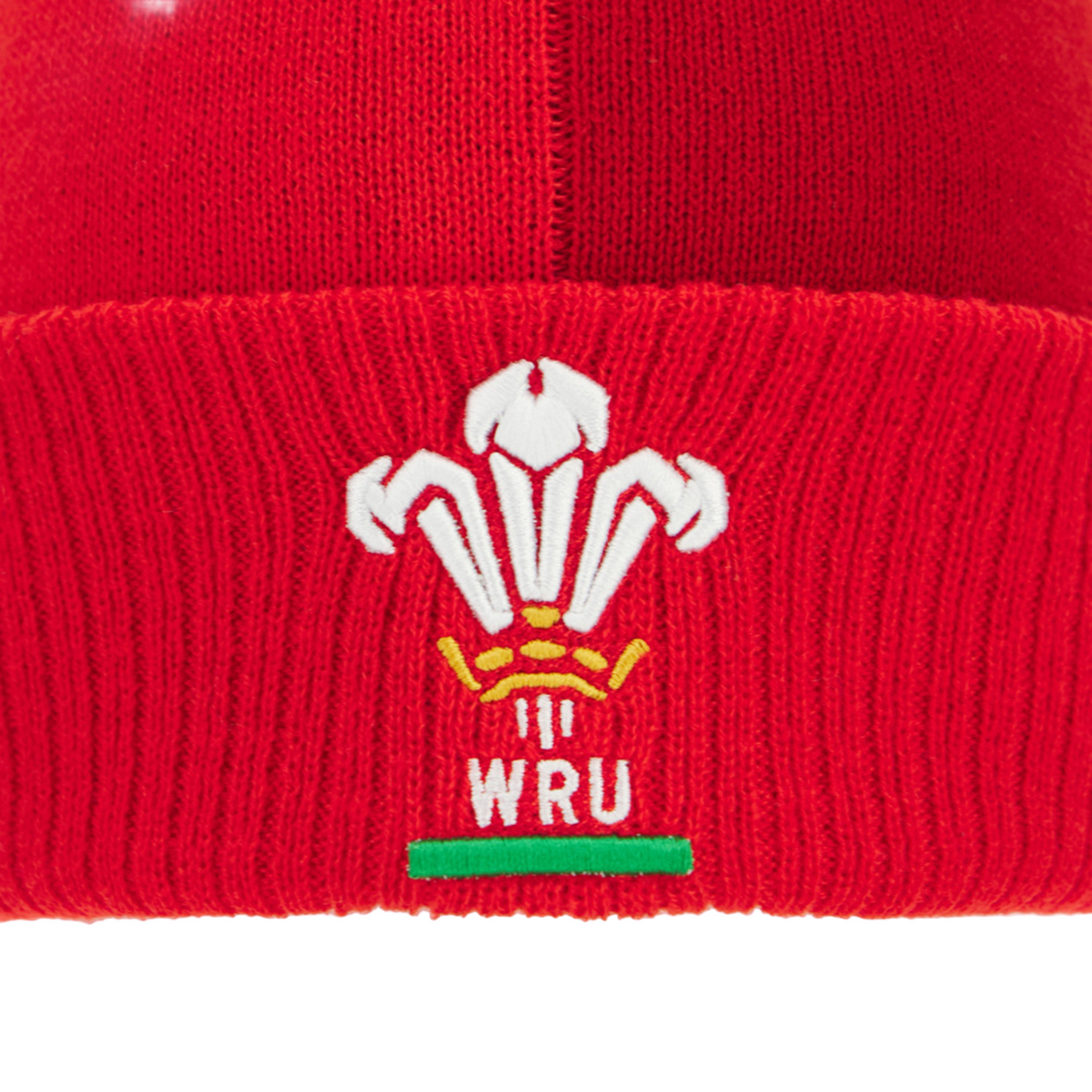 A close-up of the red Wales 25/26 Pom-Pom Beanie by Macron, featuring the Welsh Rugby Union logo embroidered in white, yellow, and green.