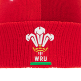 A close-up of the red Wales 25/26 Pom-Pom Beanie by Macron, featuring the Welsh Rugby Union logo embroidered in white, yellow, and green.
