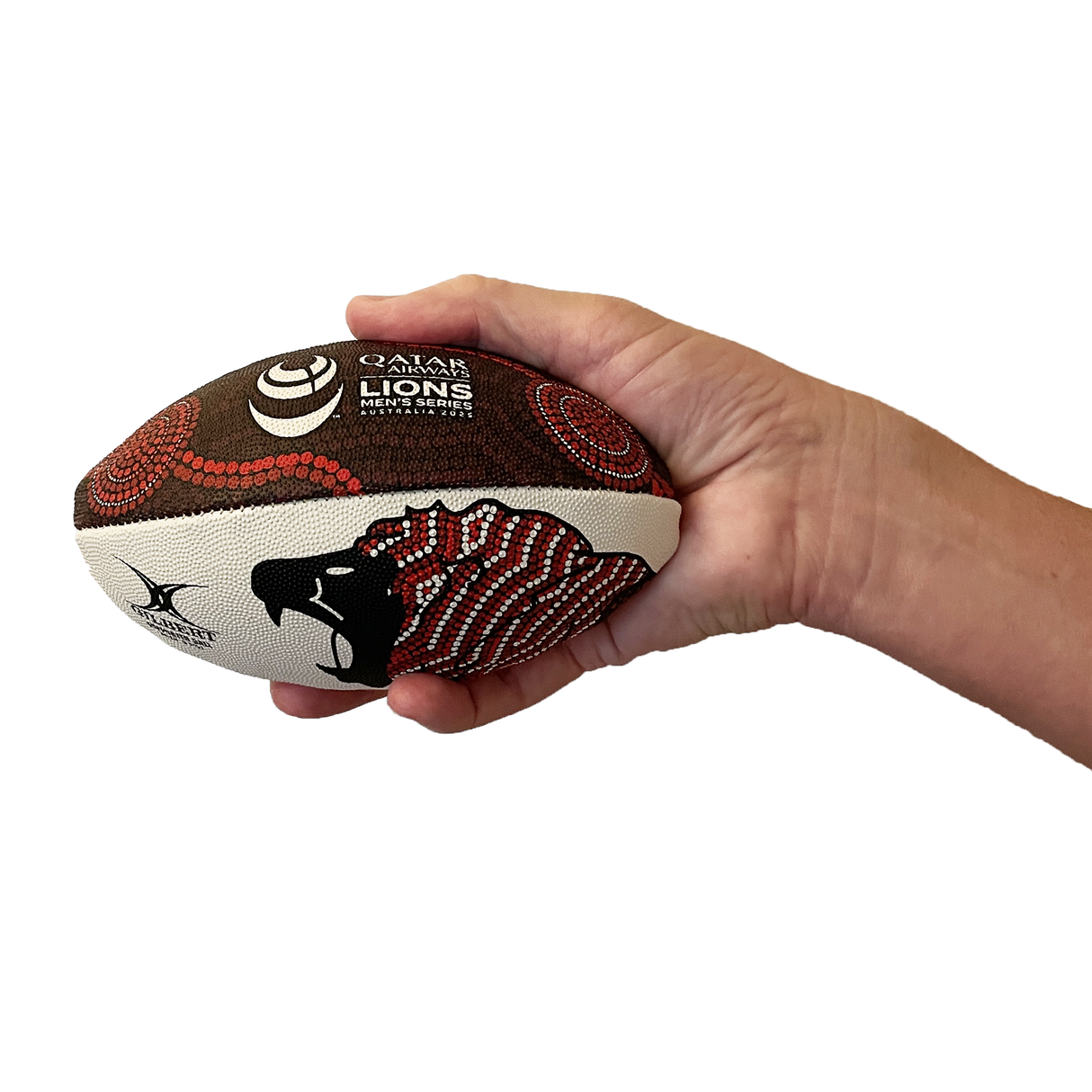 A hand holds the Gilbert British and Irish Lions Series 2025 Supporter Mini Ball, featuring Aboriginal-inspired art with a lion’s head and red dot patterns, against a plain white background.