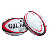Displayed are two Gilbert Orion Match Balls in white, red, and black colors with prominent “GILBERT” branding—one standing upright and the other lying on its side. These high-performance rugby balls feature the advanced Truflight valve for enhanced accuracy and flight stability.