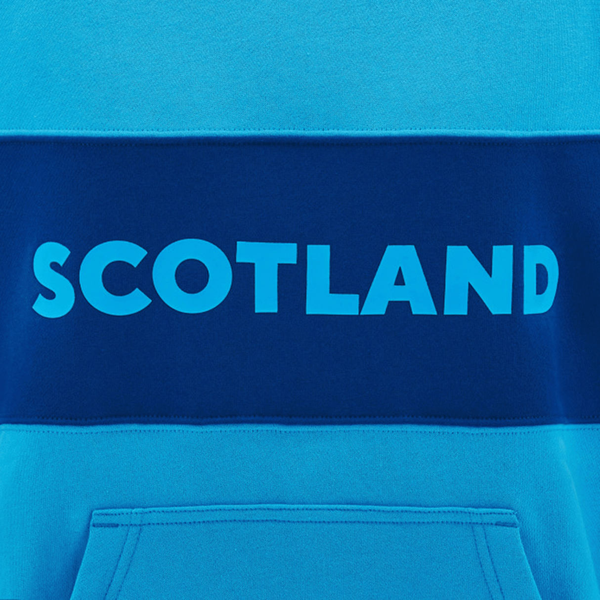 Close-up of the Scotland Block Leisure Hoodie 24/25 by Macron, featuring the word "Scotland" in bold, light blue letters on a darker blue horizontal stripe across the chest. A front pocket is visible at the bottom.