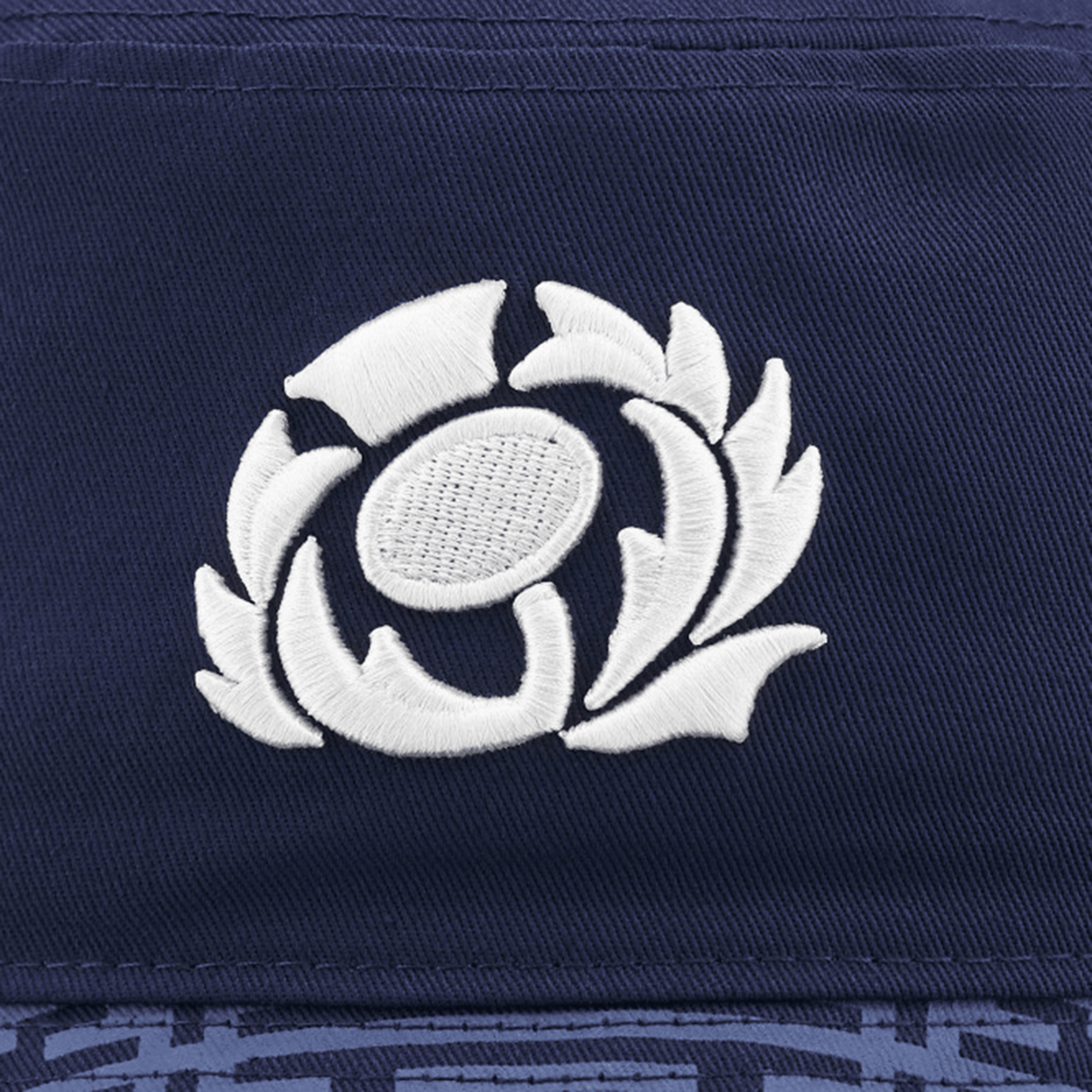 Close-up of a navy blue fabric featuring an embroidered white emblem of a stylized floral design, reminiscent of the 3D embroidered Scotland crest on the Scotland Bucket Hat 24/25 by Macron.