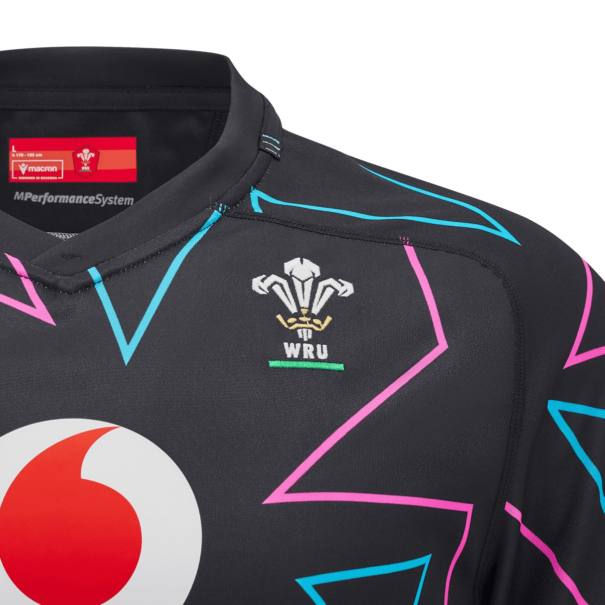 The Wales 24/25 Pathway Away Jersey by Macron, a black rugby jersey for the 24/25 season, showcases a lively geometric pattern in pink and teal. It prominently displays the Welsh Rugby Union logo and includes the Vodafone sponsor.