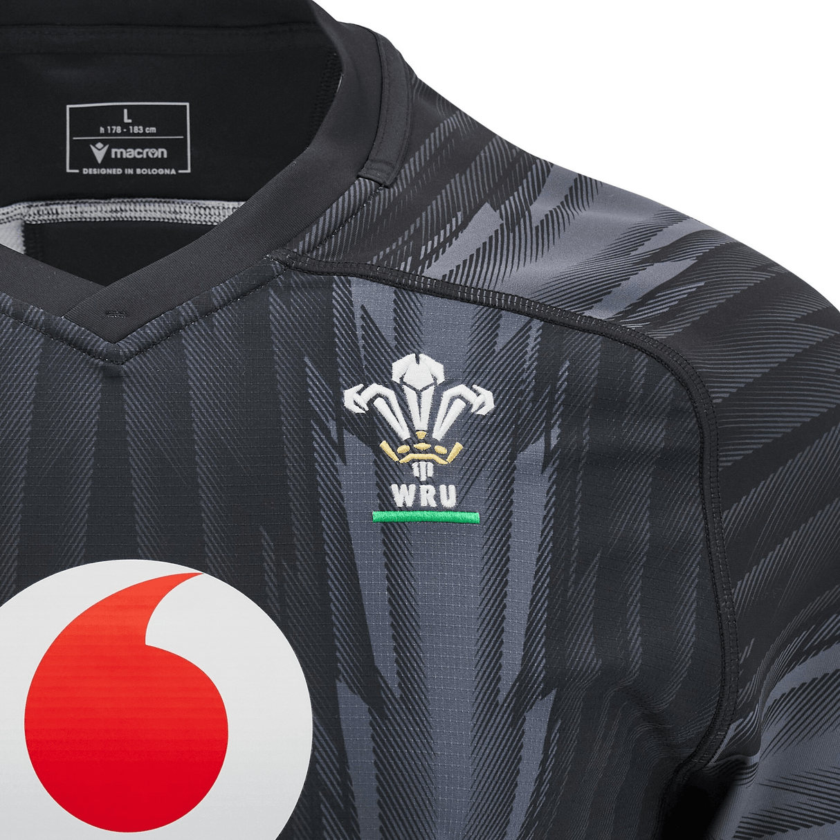 A close-up of the Wales 24/25 Training Jersey by Macron in black, displaying a white and red logo along with the WRU emblem on the chest, highlighting its status as an official Wales Training Jersey. A size "L" label is neatly positioned at the collar.