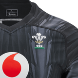 A close-up of the Wales 24/25 Training Jersey by Macron in black, displaying a white and red logo along with the WRU emblem on the chest, highlighting its status as an official Wales Training Jersey. A size "L" label is neatly positioned at the collar.
