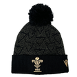 Introducing the Wales 24/25 Black Pom Pom Beanie by Macron: A stylish black knit beanie adorned with the iconic Wales crest and a playful pom-pom, enhanced by a patterned design. Perfect for showing your support, this accessory is both cozy and fashionable.