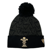 Introducing the Wales 24/25 Black Pom Pom Beanie by Macron: A stylish black knit beanie adorned with the iconic Wales crest and a playful pom-pom, enhanced by a patterned design. Perfect for showing your support, this accessory is both cozy and fashionable.