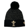 Introducing the Wales 24/25 Black Pom Pom Beanie by Macron: A stylish black knit beanie adorned with the iconic Wales crest and a playful pom-pom, enhanced by a patterned design. Perfect for showing your support, this accessory is both cozy and fashionable.